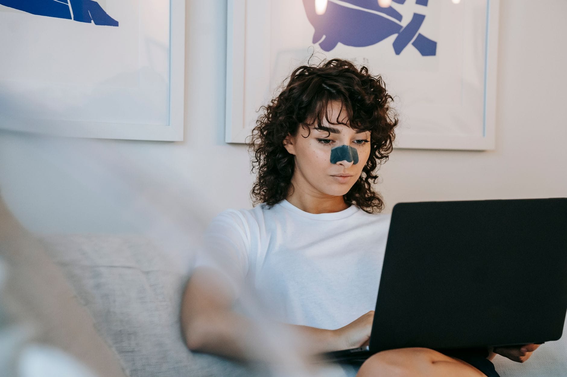 Młoda kobieta z plastrem na nosie pracuje zdalnie na laptopie na sofie